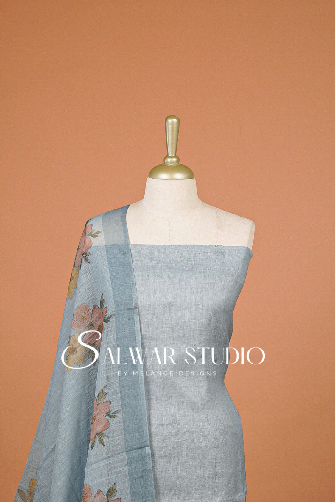 A light blue linen salwar kameez set displayed on a mannequin against a solid terracotta background. The set includes a light blue linen kurta with a square neckline and a matching light blue dupatta featuring a floral border print.