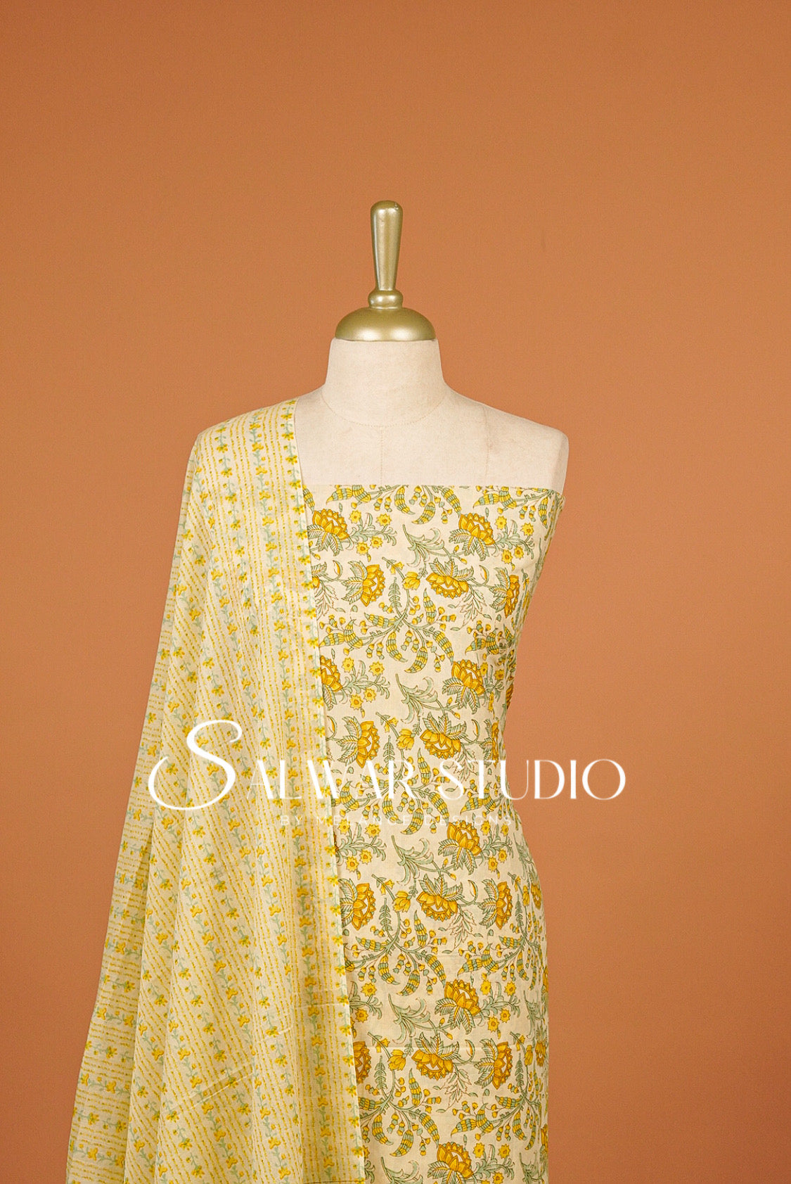 A cream-colored salwar suit set with yellow and green floral embroidery and a matching dupatta, displayed on a mannequin against a solid terracotta.