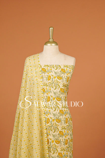 A cream-colored salwar suit set with yellow and green floral embroidery and a matching dupatta, displayed on a mannequin against a solid terracotta.