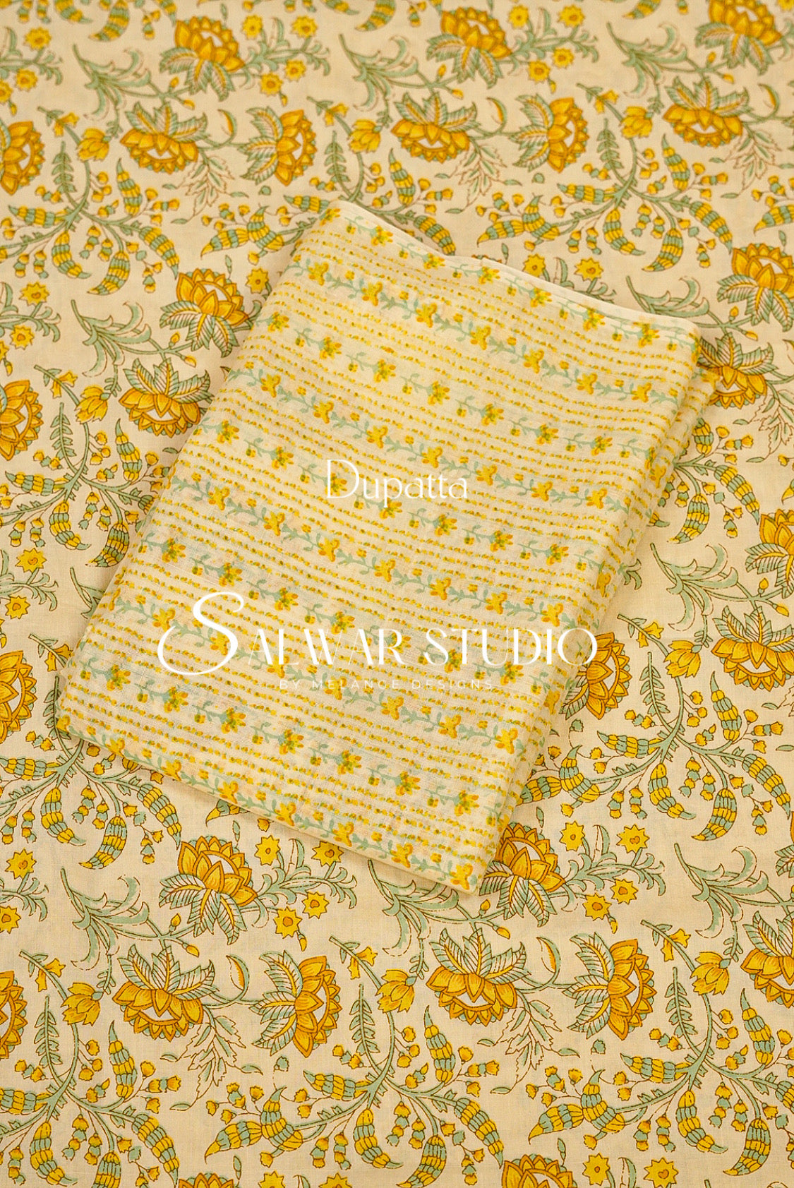 A folded piece of yellow and beige patterned fabric with green floral details, laid diagonally on top of a larger matching fabric sample. 