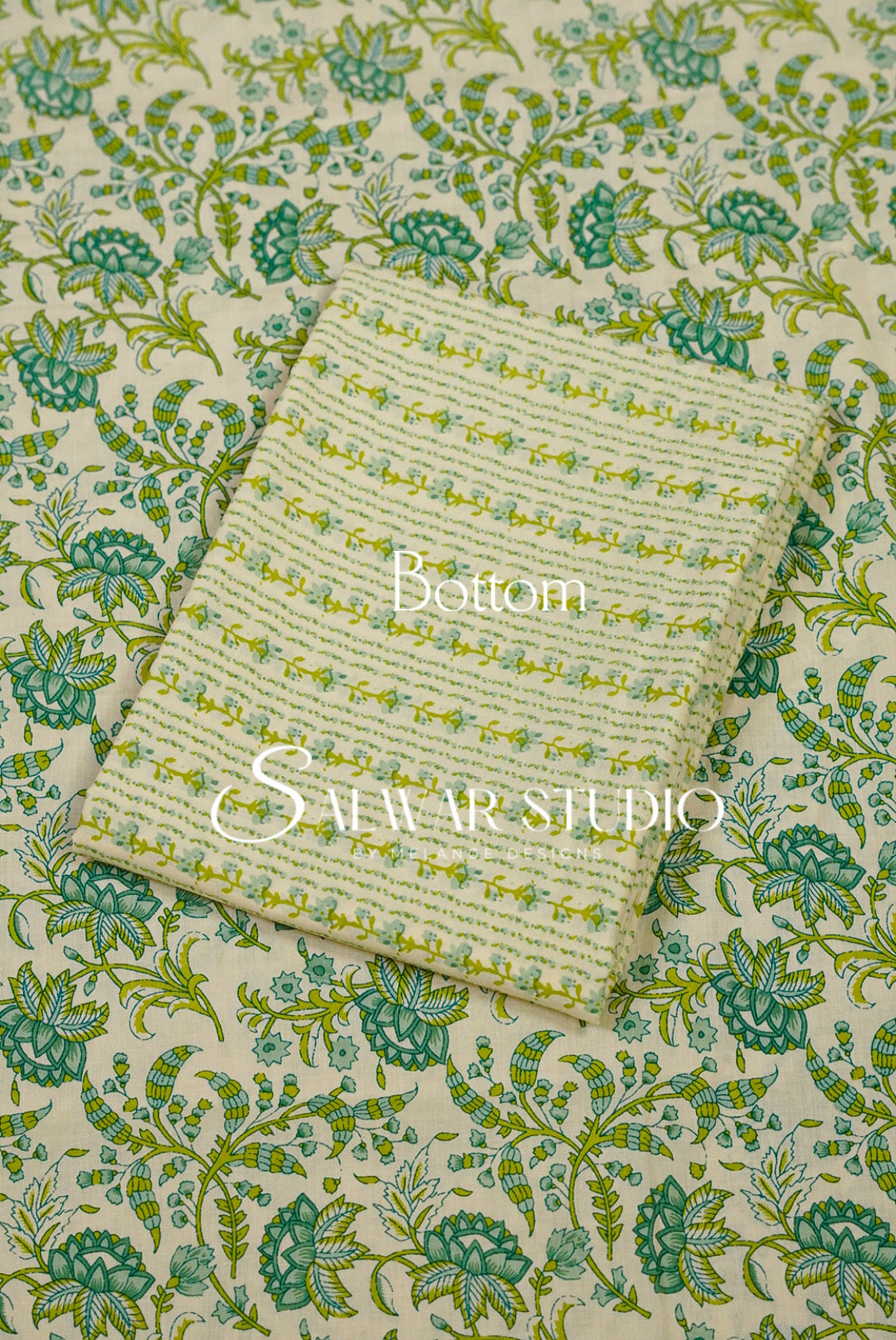 A folded cream-colored fabric napkin or small cloth with a green and yellow floral pattern, placed on top of a larger matching fabric sheet with a green and blue floral print. 
