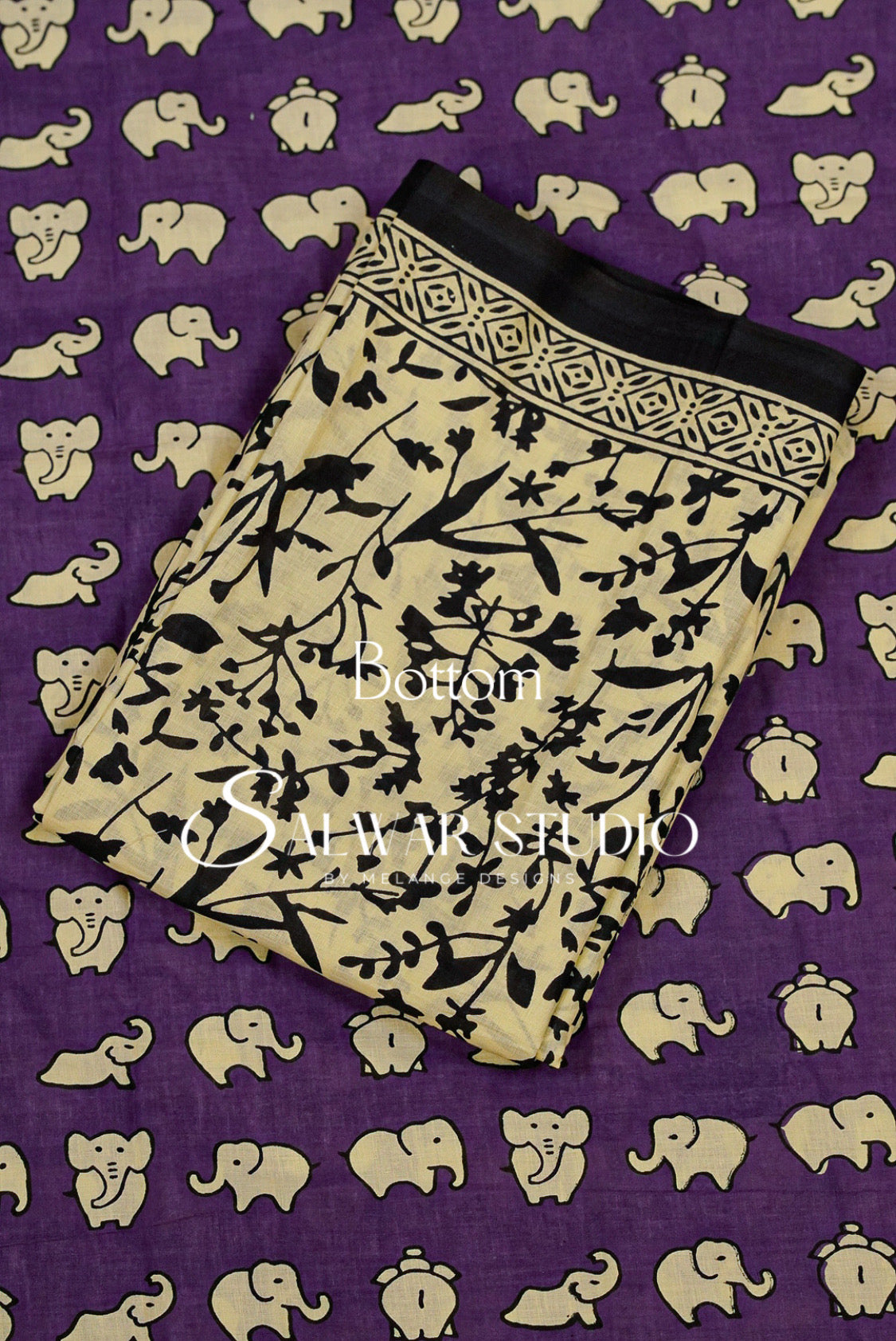 A folded beige fabric piece with a black floral and branch block print, laid diagonally on a purple background patterned with beige elephant silhouettes. 