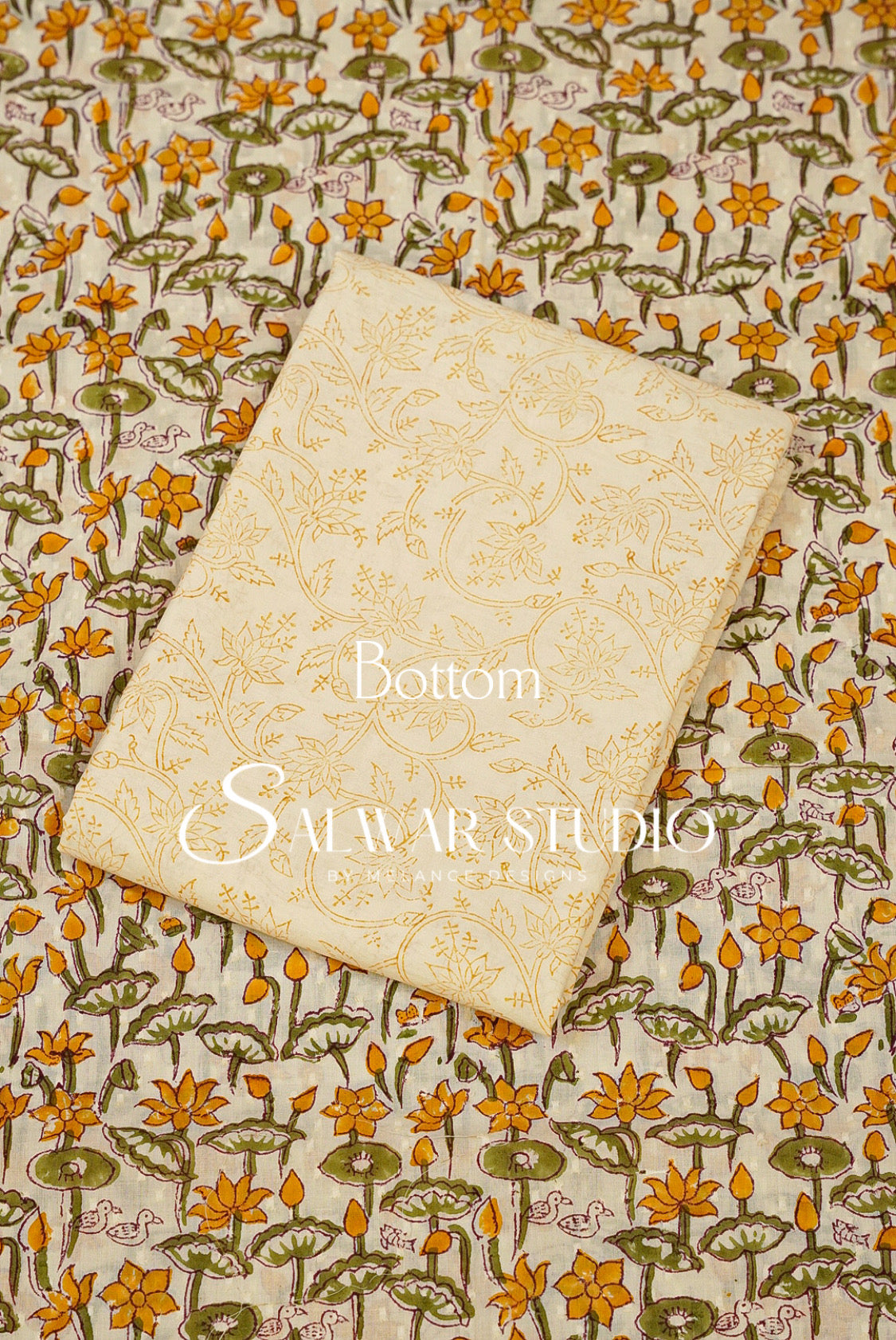 A folded cream-colored fabric piece with a subtle gold floral pattern, placed diagonally on top of a larger fabric sample featuring a dense yellow, green, and brown floral and lotus print.