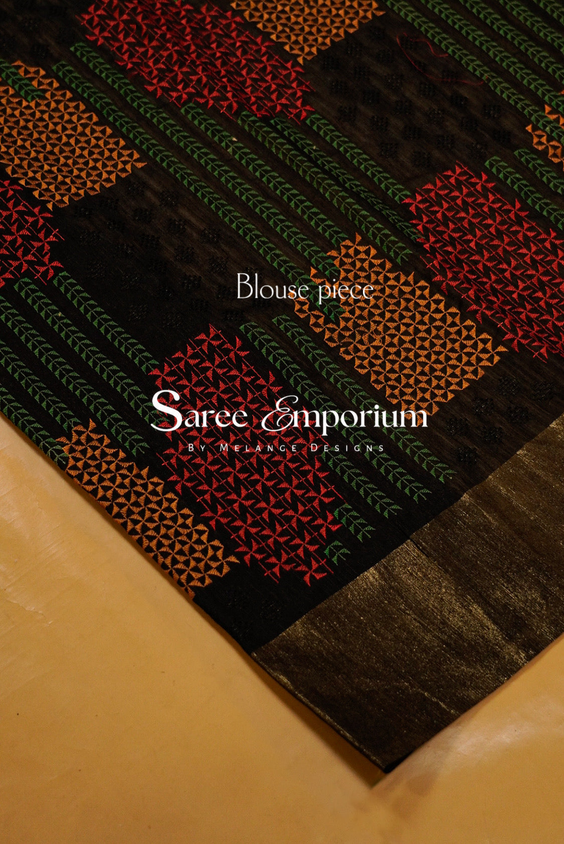 A close-up of a patterned blouse piece fabric by Saree Emporium, featuring a black base with red, orange, and green geometric and floral motifs.