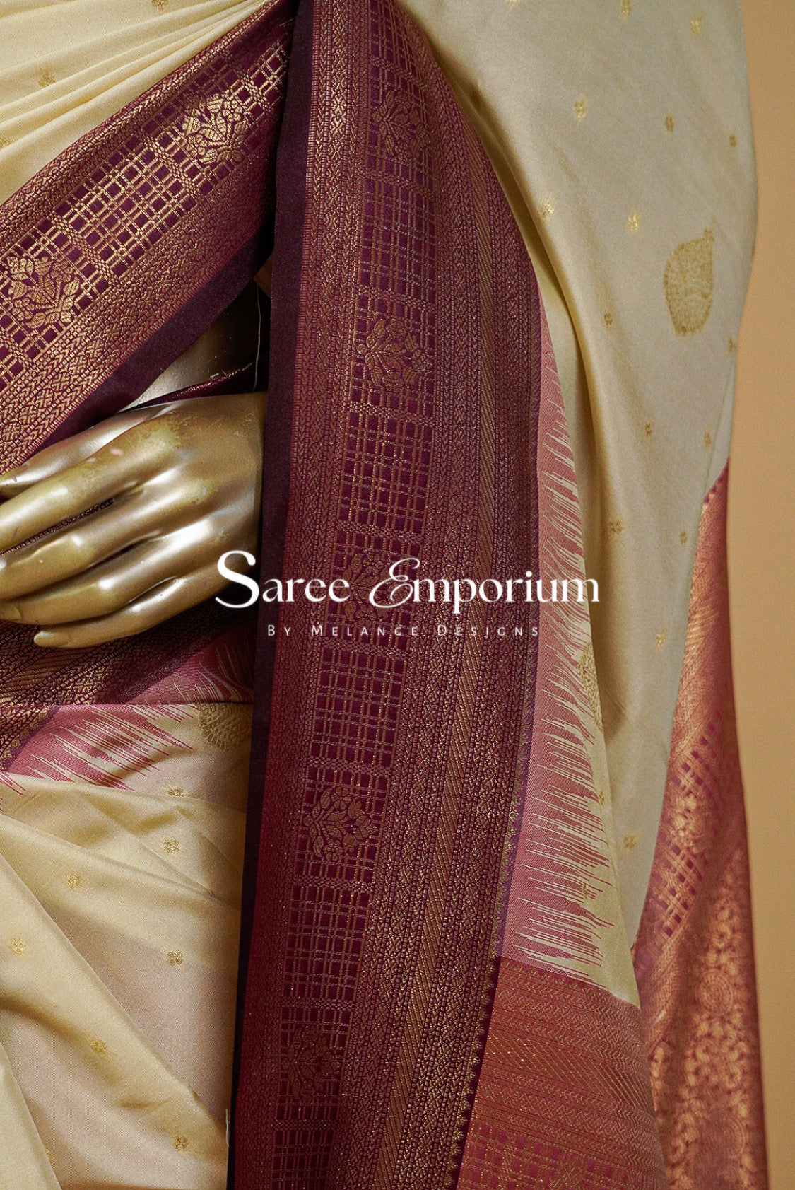 A close-up of a draped saree with a cream-colored body, deep maroon border, and gold zari woven patterns.