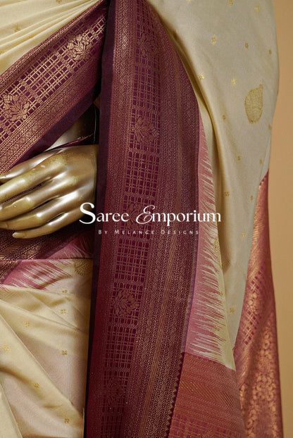 A close-up of a draped saree with a cream-colored body, deep maroon border, and gold zari woven patterns.