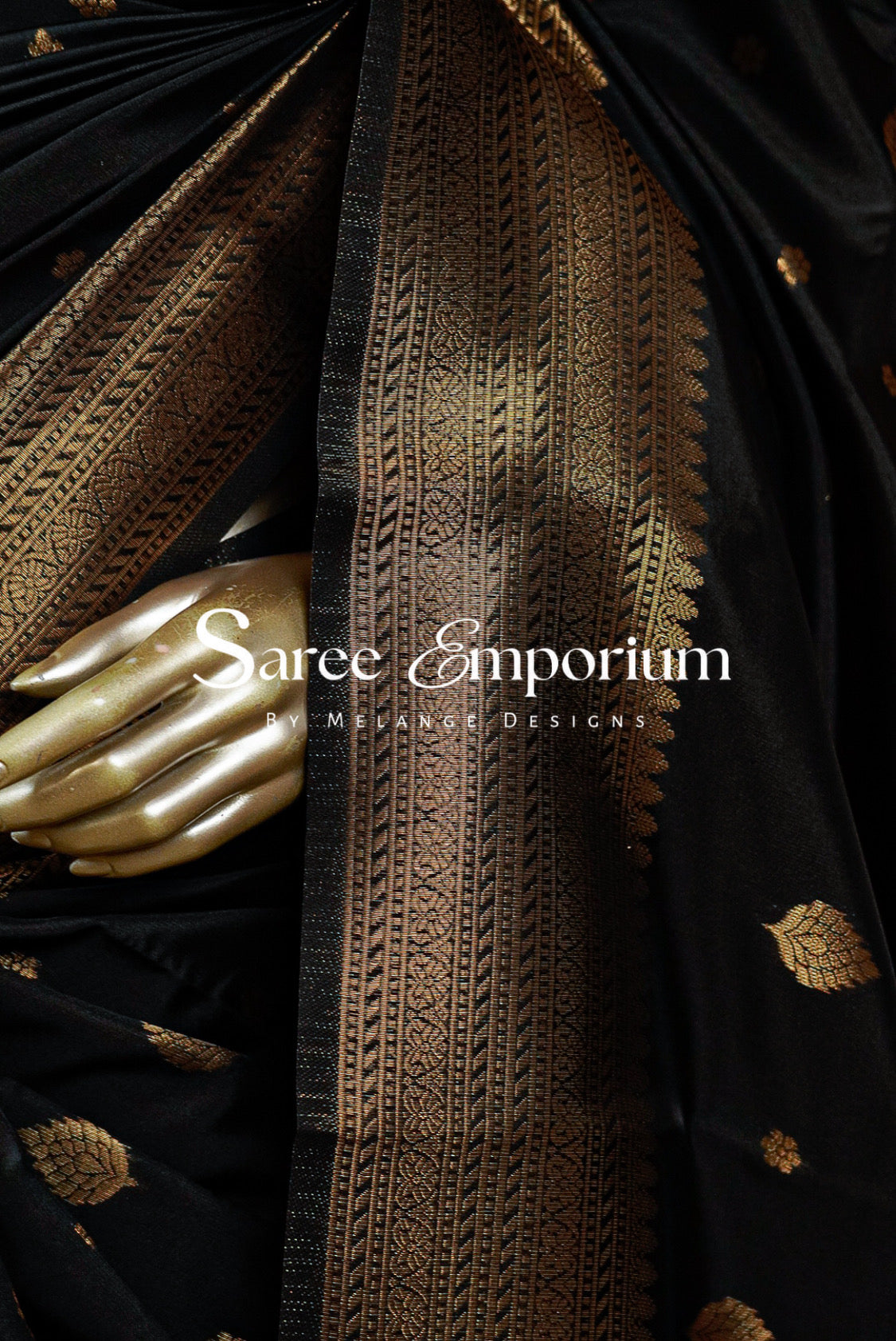 A close-up of a black silk saree with an ornate gold zari border and scattered gold motifs, draped elegantly with a metallic gold hand resting on top. 