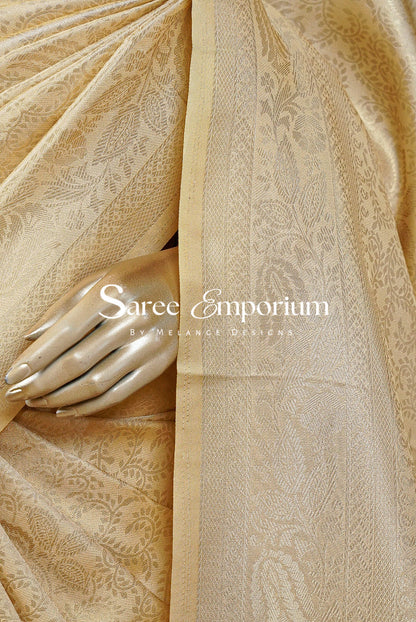 A close-up of a beige silk saree with a subtle floral jacquard pattern and a matching border, draped elegantly with a metallic gold mannequin hand resting on it.