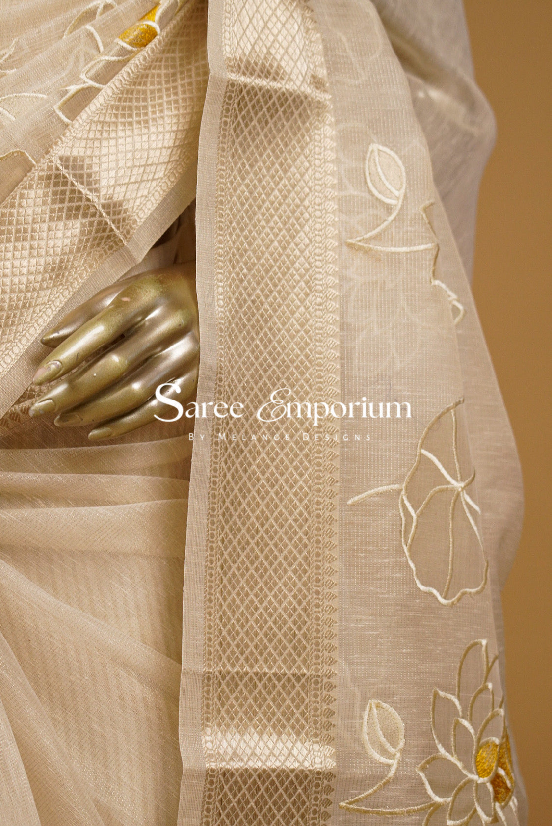 A close-up of a beige silk saree draped elegantly, featuring a wide border with geometric woven patterns and embroidered floral motifs in beige and gold. 
