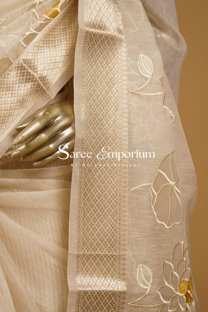 A close-up of a beige silk saree draped elegantly, featuring a wide border with geometric woven patterns and embroidered floral motifs in beige and gold. 