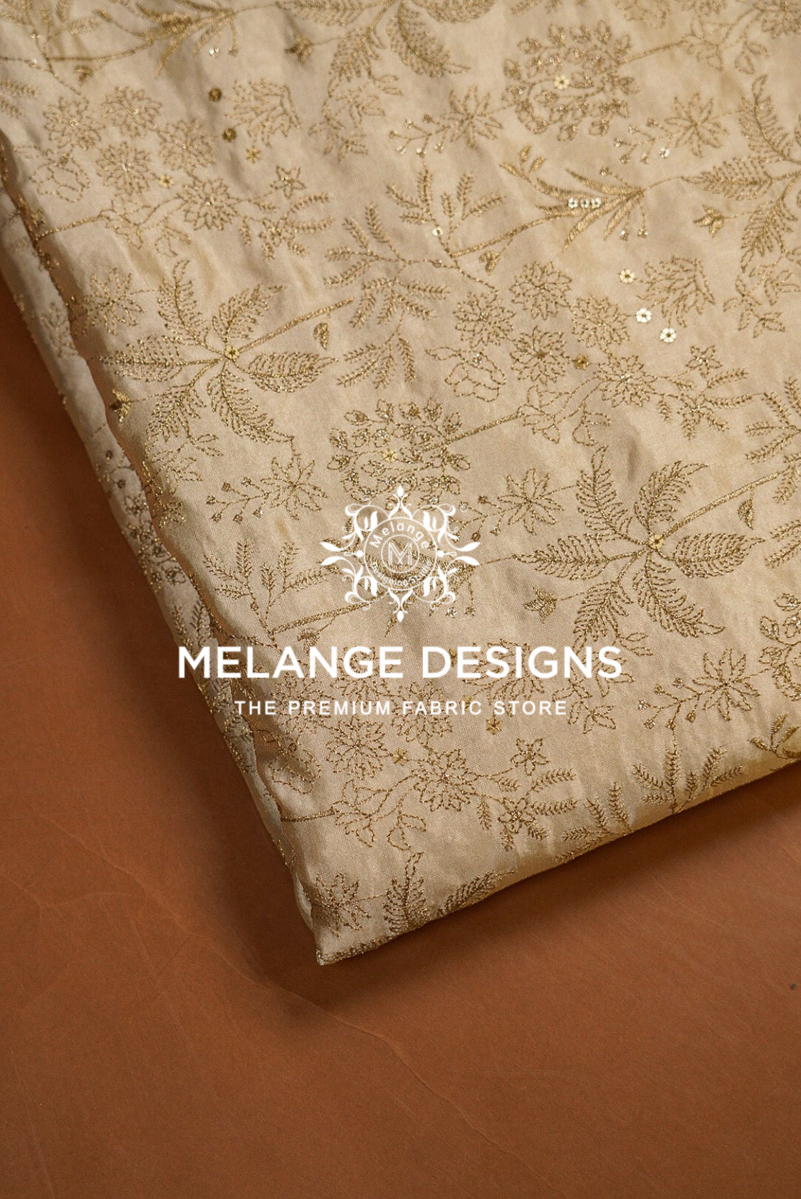 A close-up of a folded beige Tissue Satin Embroidery Fabric Dyeable with intricate gold floral embroidery and sequins, displayed on a brown surface. 