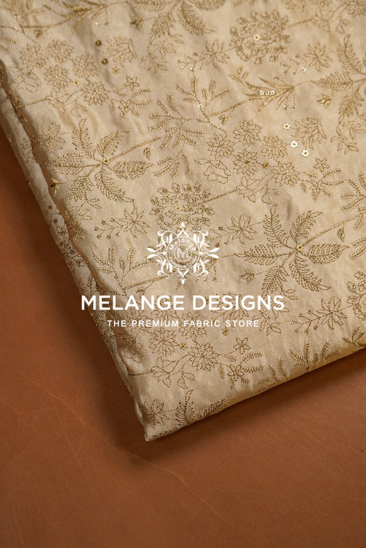 A close-up of a folded beige Tissue Satin Embroidery Fabric Dyeable with intricate gold floral embroidery and sequins, displayed on a brown surface. 