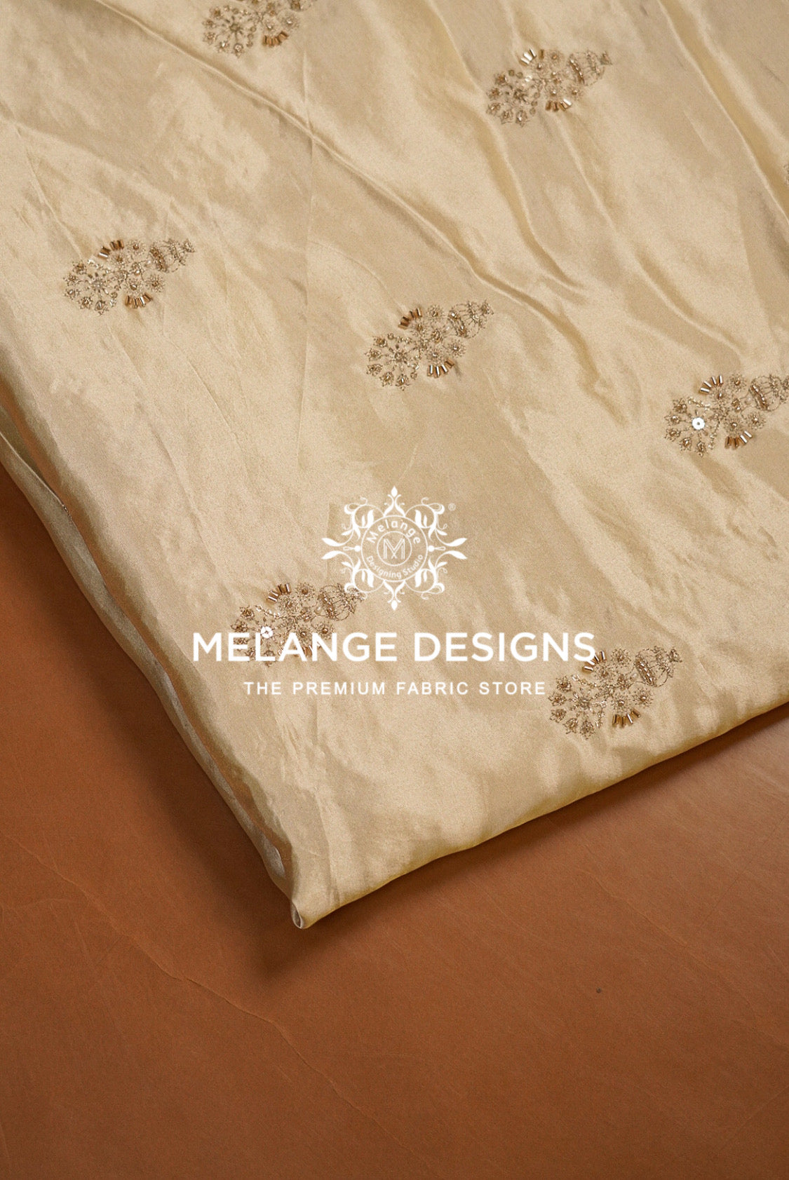 A close-up of a cream-colored Tissue Satin Embroidery Fabric Dyeable  with delicate gold floral embroidery, draped diagonally over a brown surface.