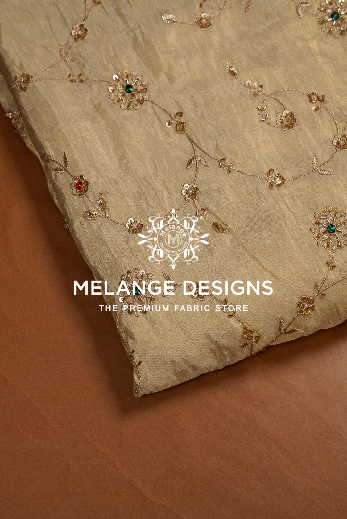 A close-up of a beige Tissue Silk Embroidery Fabric with delicate gold floral embroidery and small red and green sequin accents, laid diagonally on a brown surface. 