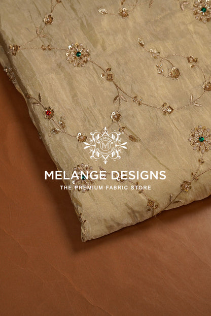 A close-up of a beige Tissue Silk Embroidery Fabric with delicate gold floral embroidery and small red and green sequin accents, laid diagonally on a brown surface. 