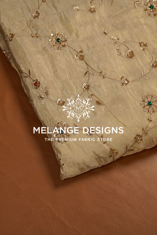 A close-up of a beige Tissue Silk Embroidery Fabric with delicate gold floral embroidery and small red and green sequin accents, laid diagonally on a brown surface. 