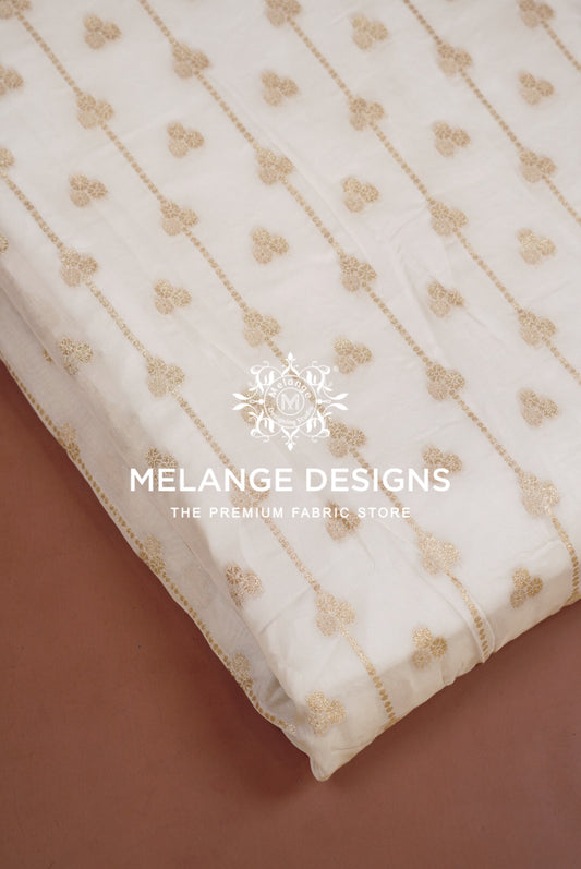 An off-white Viscose Jaqured Dyeable Fabric with delicate gold floral embroidery and vertical gold stripes, folded diagonally on a brown surface. 