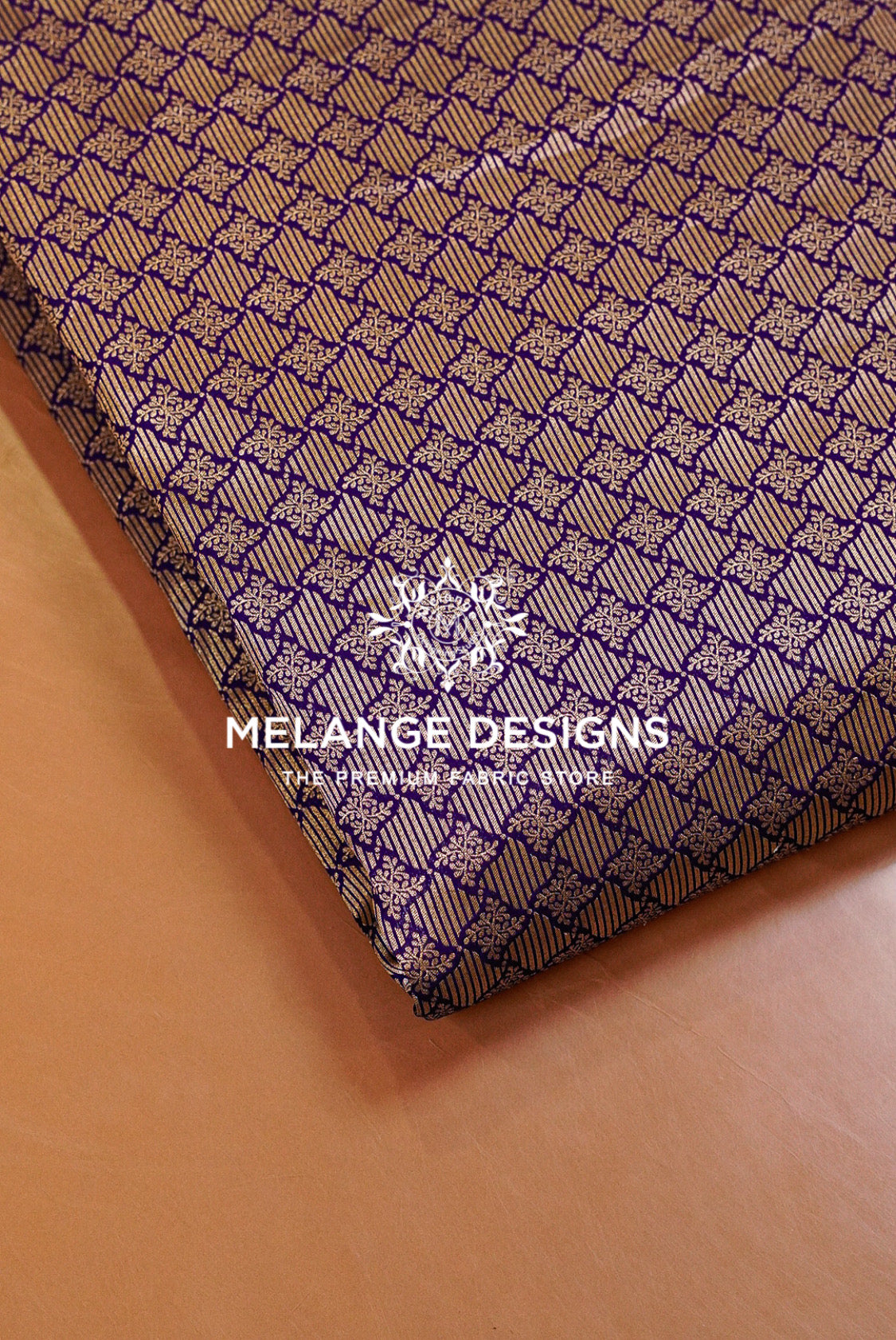 A close-up of a folded Banarasi Brocade Fabric or textile sample with a repeating geometric pattern in purple and beige, placed on a tan surface. 