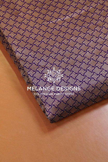 A close-up of a folded Banarasi Brocade Fabric or textile sample with a repeating geometric pattern in purple and beige, placed on a tan surface. 