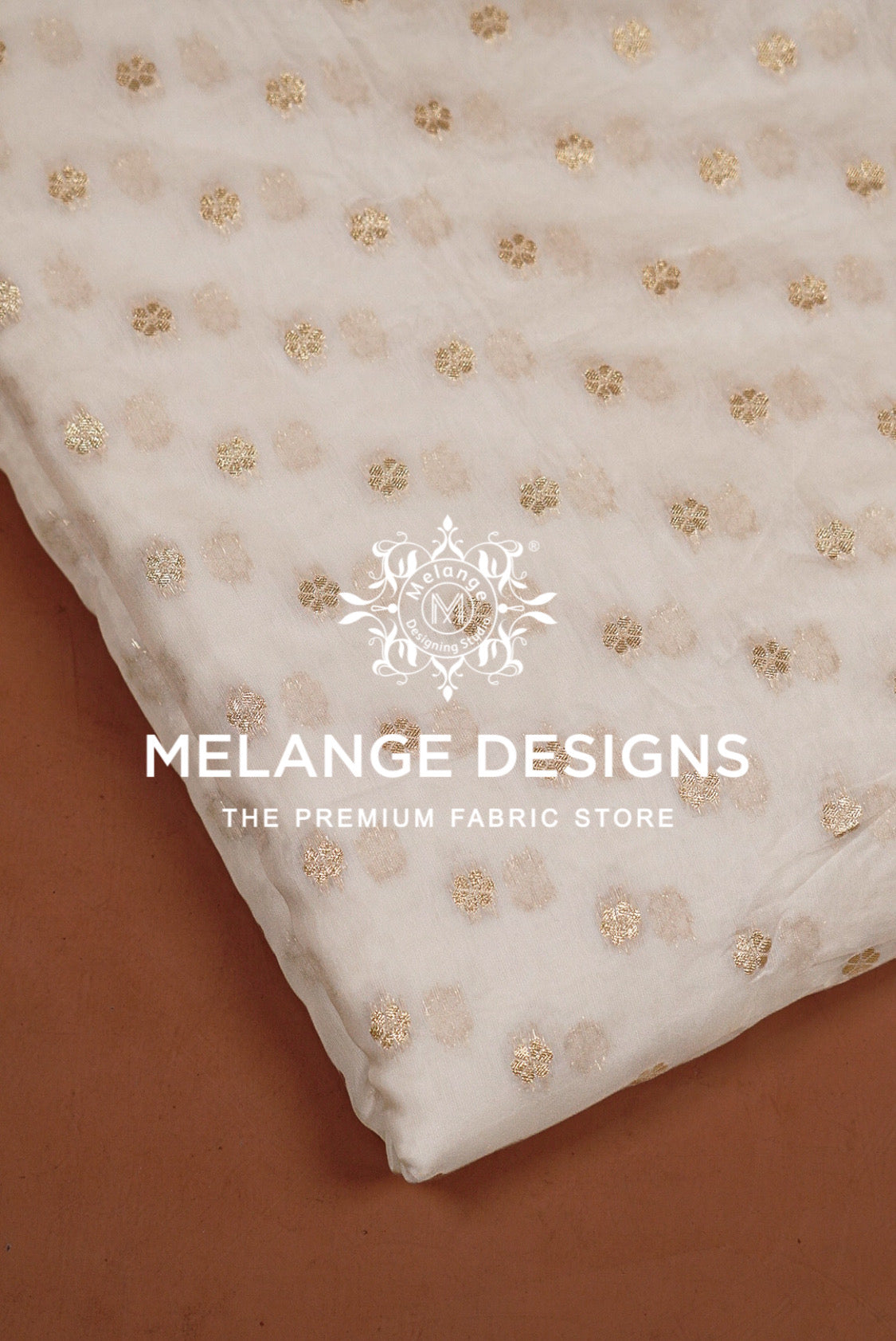 A close-up of a white or off-white Cotton Chanderi Brocade Dyeable  Fabric with small scattered gold floral or dot motifs, laid diagonally over a warm brown surface.