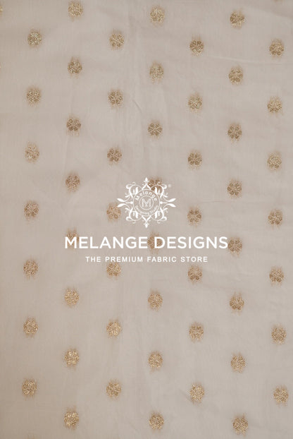 A close-up of a light beige Cotton Chanderi Brocade Dyeable  Fabric with a repeating pattern of small, textured gold floral or starburst motifs.