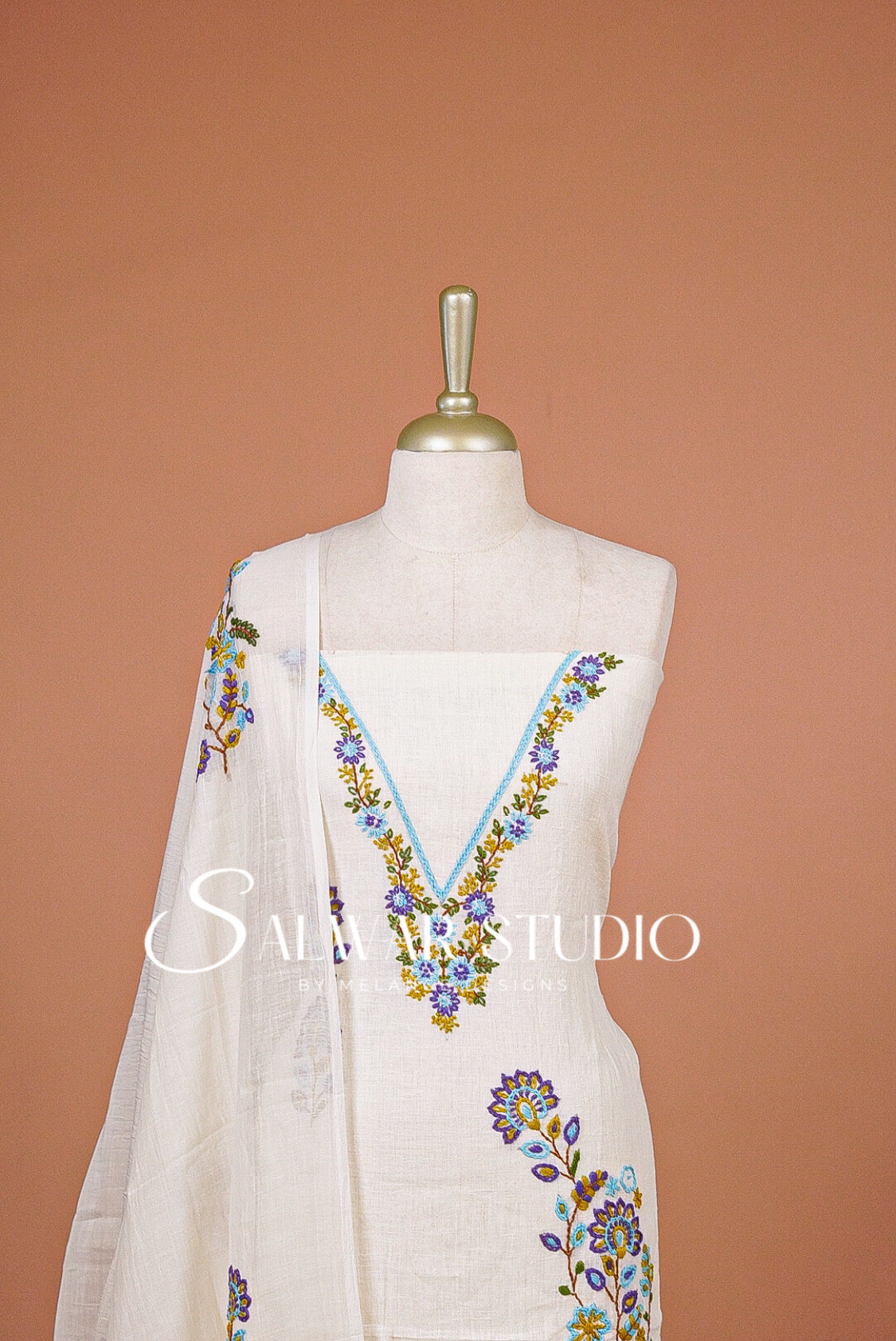 An off-white cotton kurta set with a matching dupatta draped over a mannequin, featuring colorful floral embroidery in blue, purple, and yellow around the V-neckline and on the dupatta. 