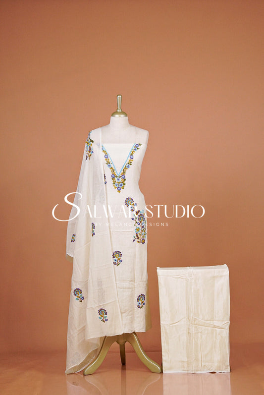 A white traditional outfit displayed on a mannequin against a warm peach studio backdrop, with a matching white dupatta draped over the shoulders and a folded matching garment beside the stand. The outfit has colorful floral embroidery concentrated around the neckline and scattered along the lower length.