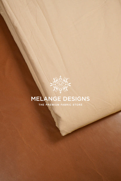 A close-up of a folded cream-colored Premium Cora Cotton Fabric Dyeable likely a bed sheet or duvet cover, resting on a brown leather surface. 