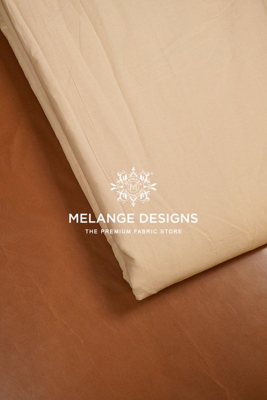 A close-up of a folded cream-colored Premium Cora Cotton Fabric Dyeable likely a bed sheet or duvet cover, resting on a brown leather surface. 