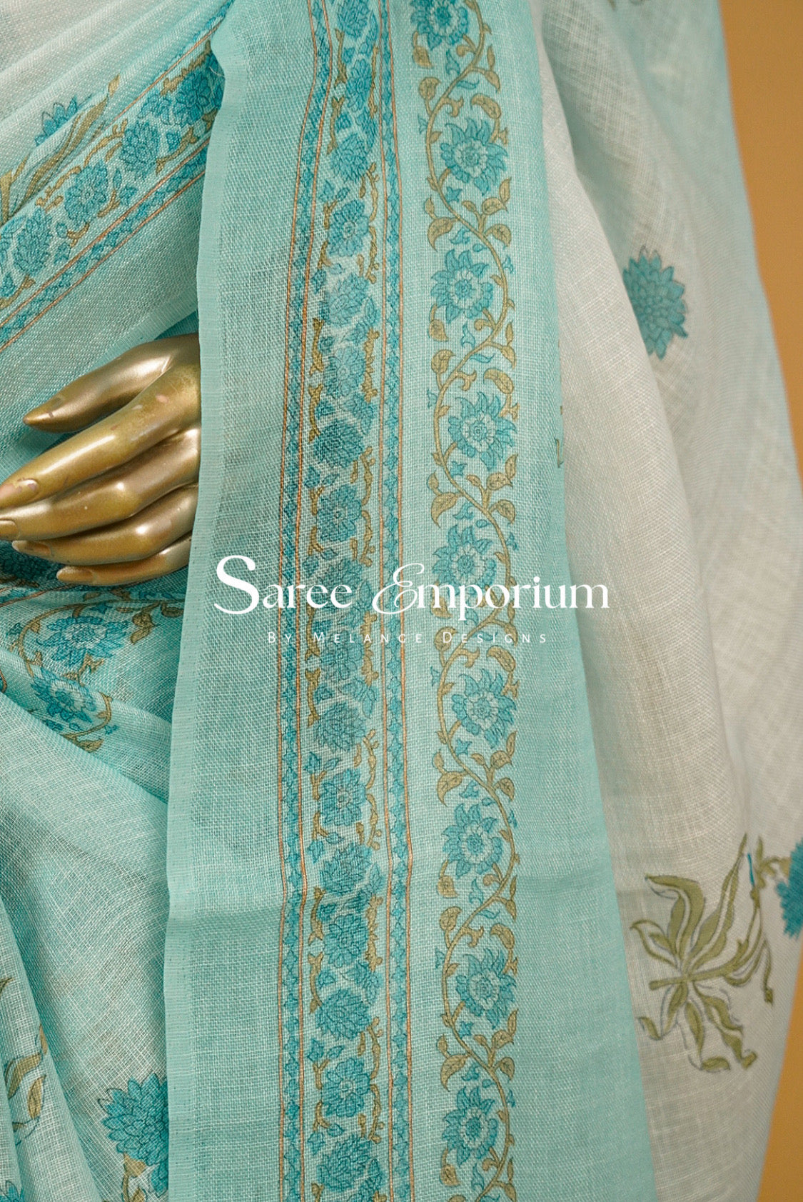 A close-up of a light aqua blue saree fabric with teal floral prints and gold border embroidery, draped over a white saree with gold floral motifs.