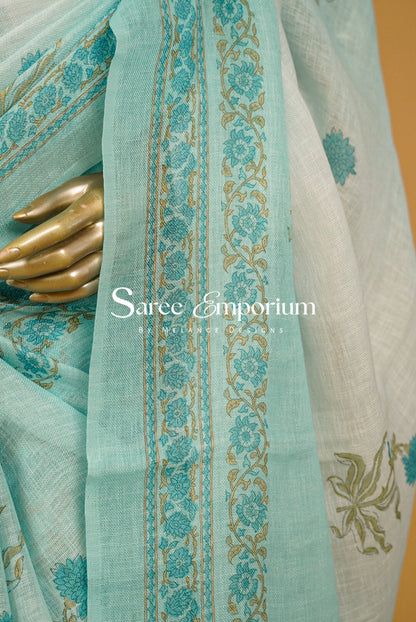 A close-up of a light aqua blue saree fabric with teal floral prints and gold border embroidery, draped over a white saree with gold floral motifs.