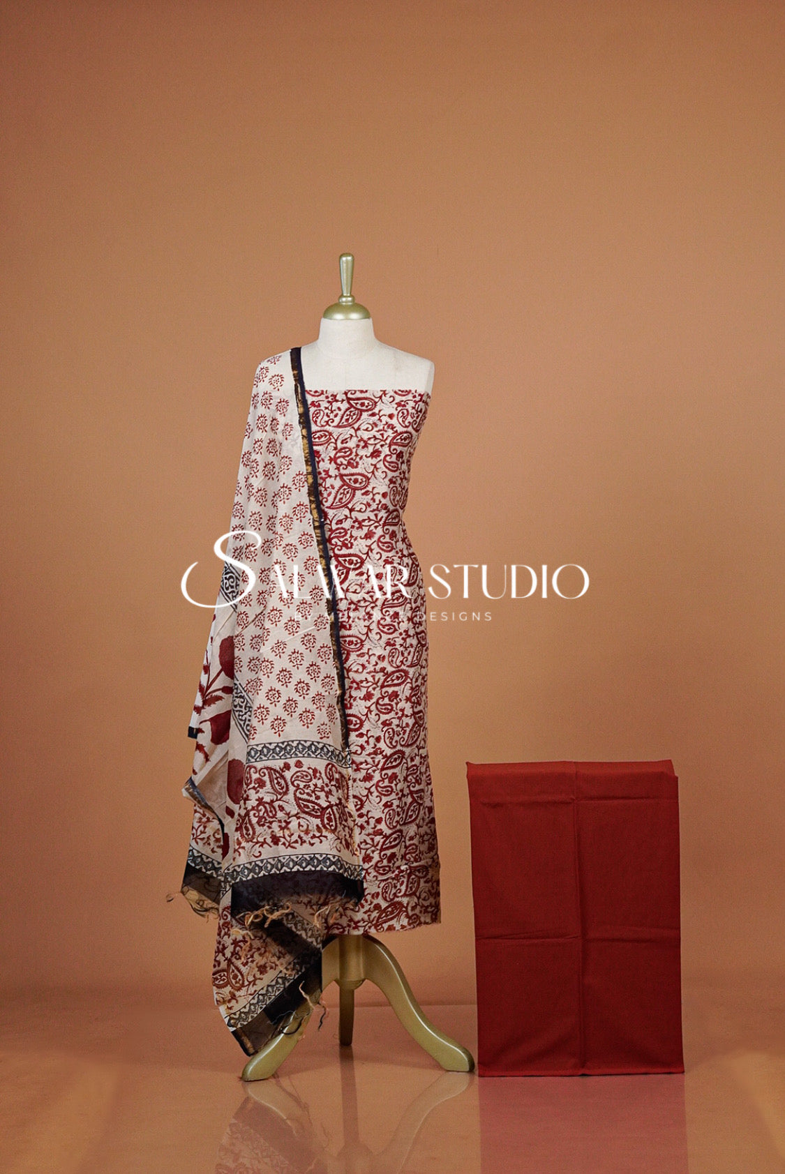 A printed dress with a matching red fabric box beside it. The dress has a red and beige floral block-print pattern with a black border, and a white dupatta with red and black motifs.