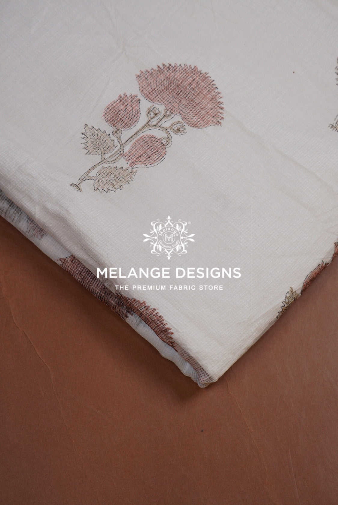 A close-up of a cream-colored Printed Cotton Cotta Fabric  with a dusty rose and beige floral block print, folded diagonally on a brown textured surface. 