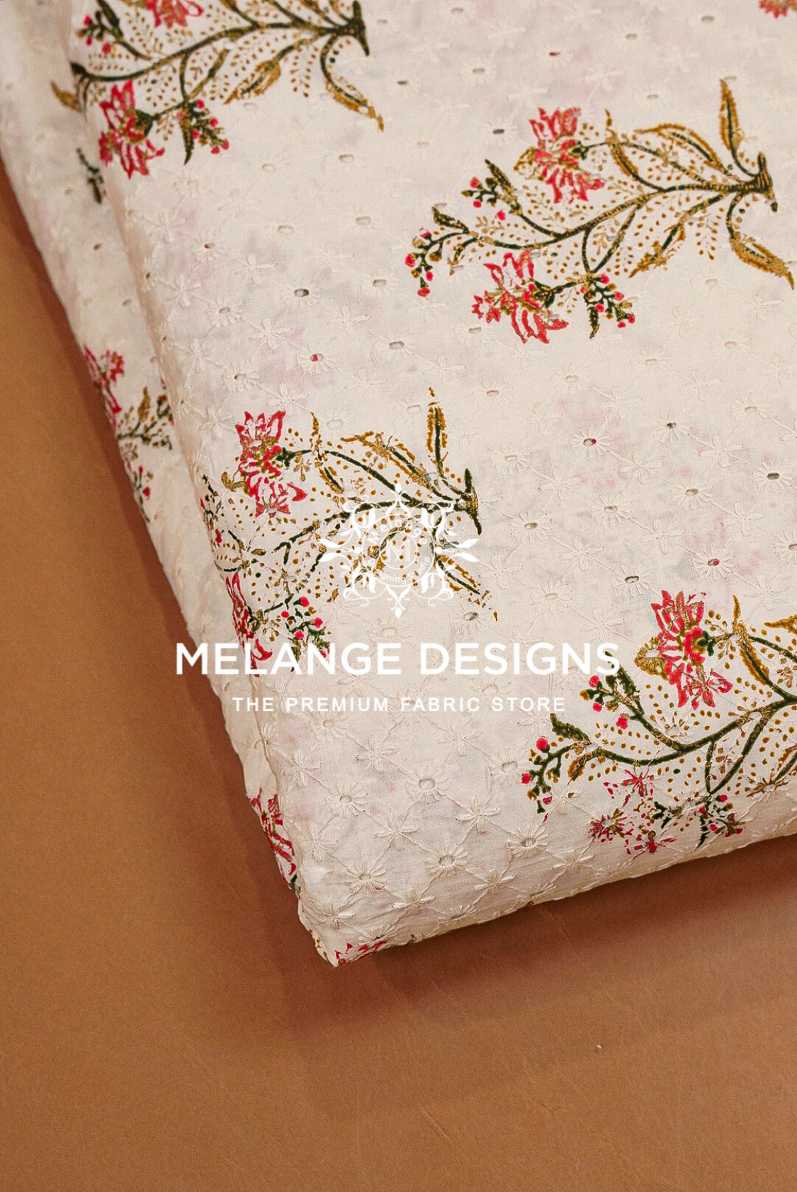 A close-up of a folded cream-colored Printed Cotton Hacoba Fabric BJ0047 with a textured floral print featuring pink flowers, green leaves, and gold accents, displayed on a brown surface. 