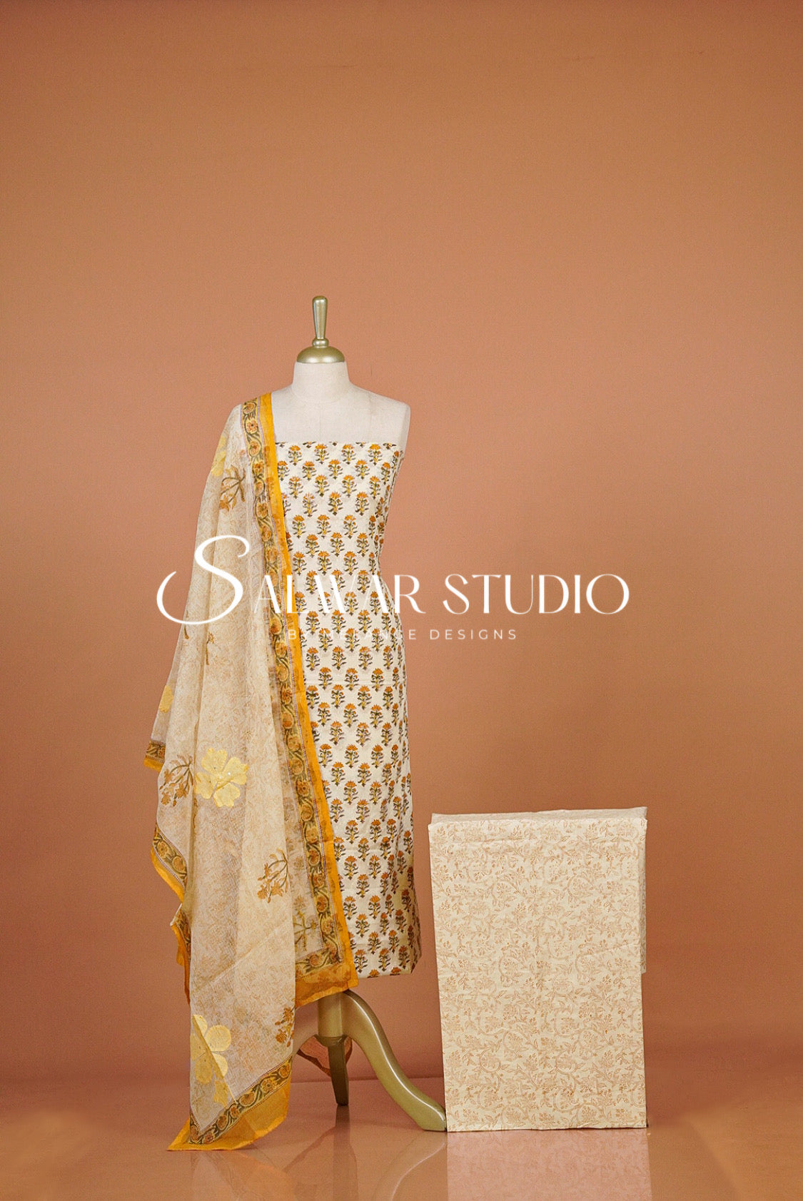 A cream-colored ethnic suit set featuring a printed kurta, matching dupatta, and a folded fabric box. 