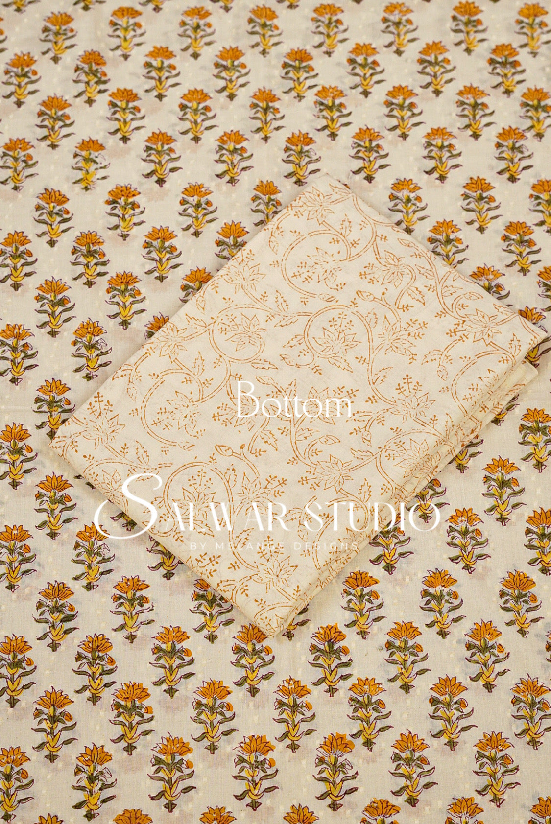 A folded cream-colored fabric piece with a gold floral pattern, placed diagonally over a larger matching fabric sheet featuring a repeating orange and brown floral block print on a cream .