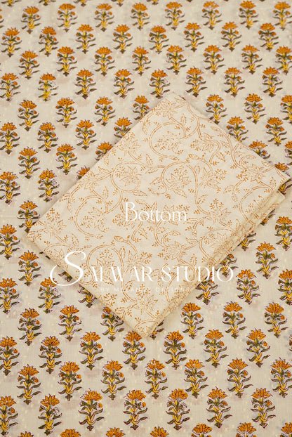 A folded cream-colored fabric piece with a gold floral pattern, placed diagonally over a larger matching fabric sheet featuring a repeating orange and brown floral block print on a cream .