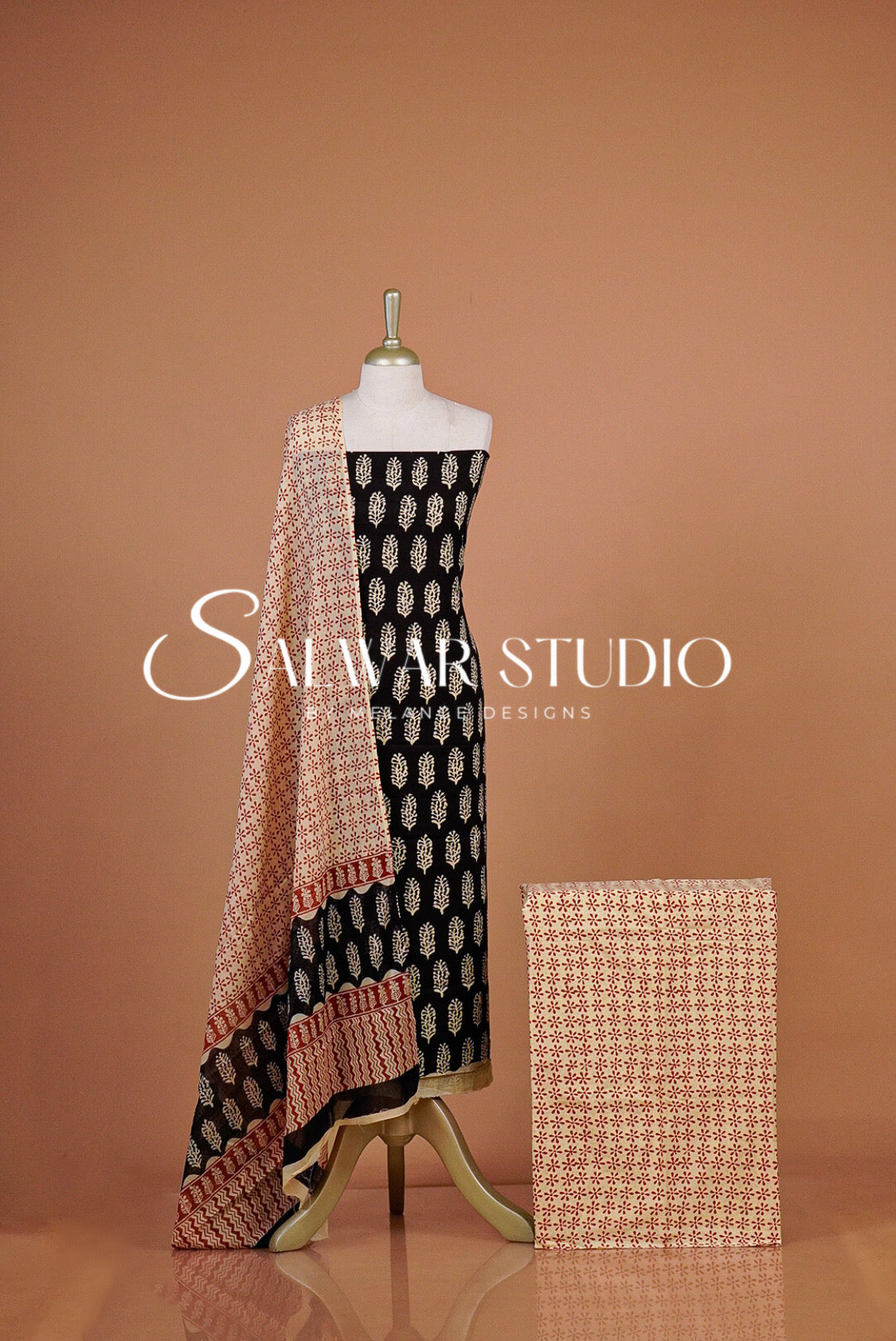A women's salwar suit set displayed on a The set includes a black salwar with beige and white motifs, a beige printed dupatta with peach border details, and a matching beige printed fabric sample.