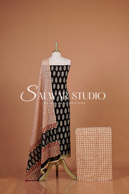 A women's salwar suit set displayed on a The set includes a black salwar with beige and white motifs, a beige printed dupatta with peach border details, and a matching beige printed fabric sample.