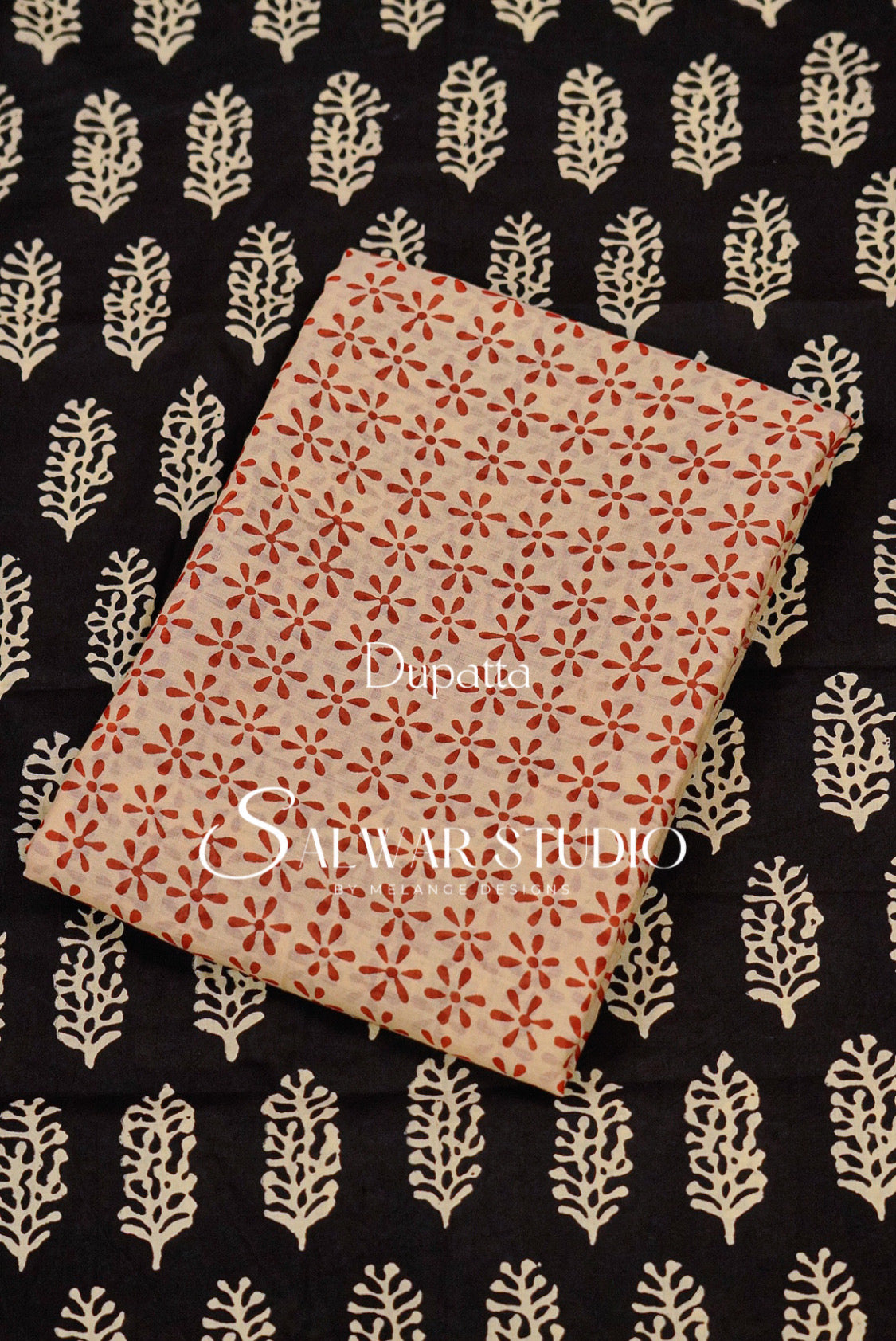 A folded piece of fabric with a red and beige block print pattern, displayed diagonally on top of a black fabric with a cream-colored leaf block print.