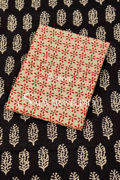 A folded fabric sample with a beige background and red floral block print, placed diagonally on top of a black fabric with a cream leaf pattern.