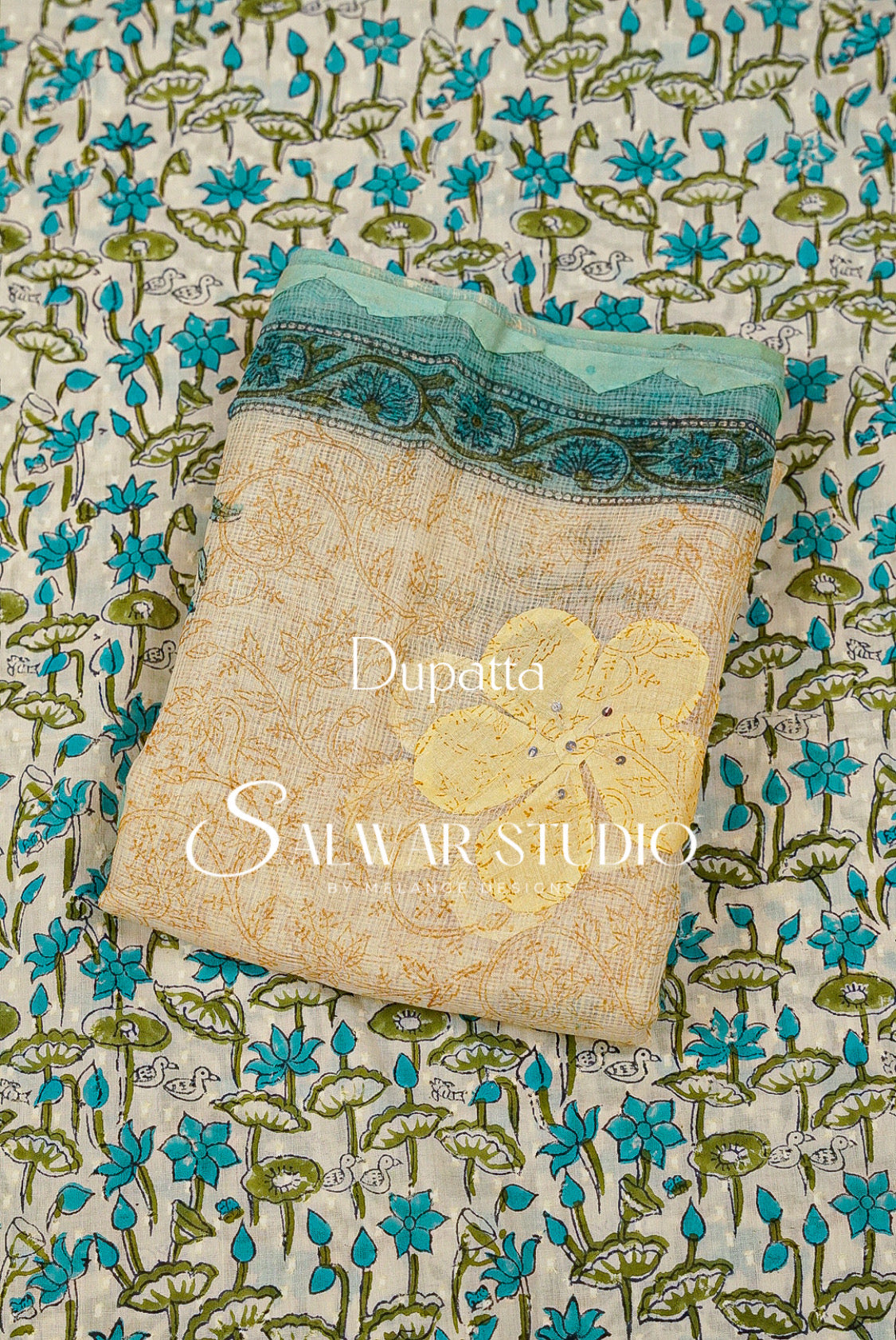 A folded dupatta fabric sample with a beige base, teal floral border, and yellow embroidered floral motif, placed on top of a matching patterned fabric .
