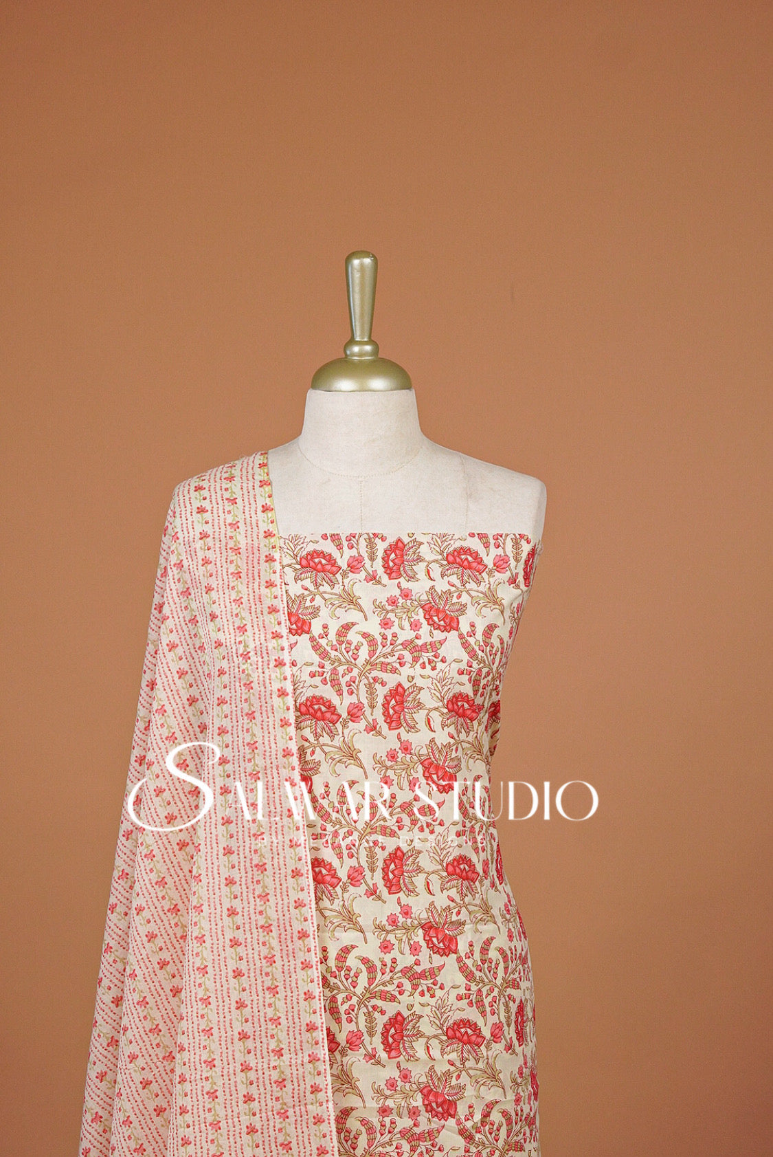 A cream-colored traditional outfit . The outfit features a red and gold floral print on the top and a matching patterned dupatta draped over the shoulder.