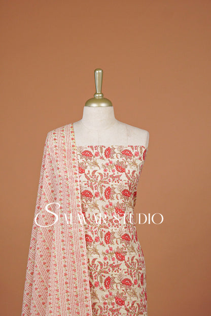 A cream-colored traditional outfit . The outfit features a red and gold floral print on the top and a matching patterned dupatta draped over the shoulder.