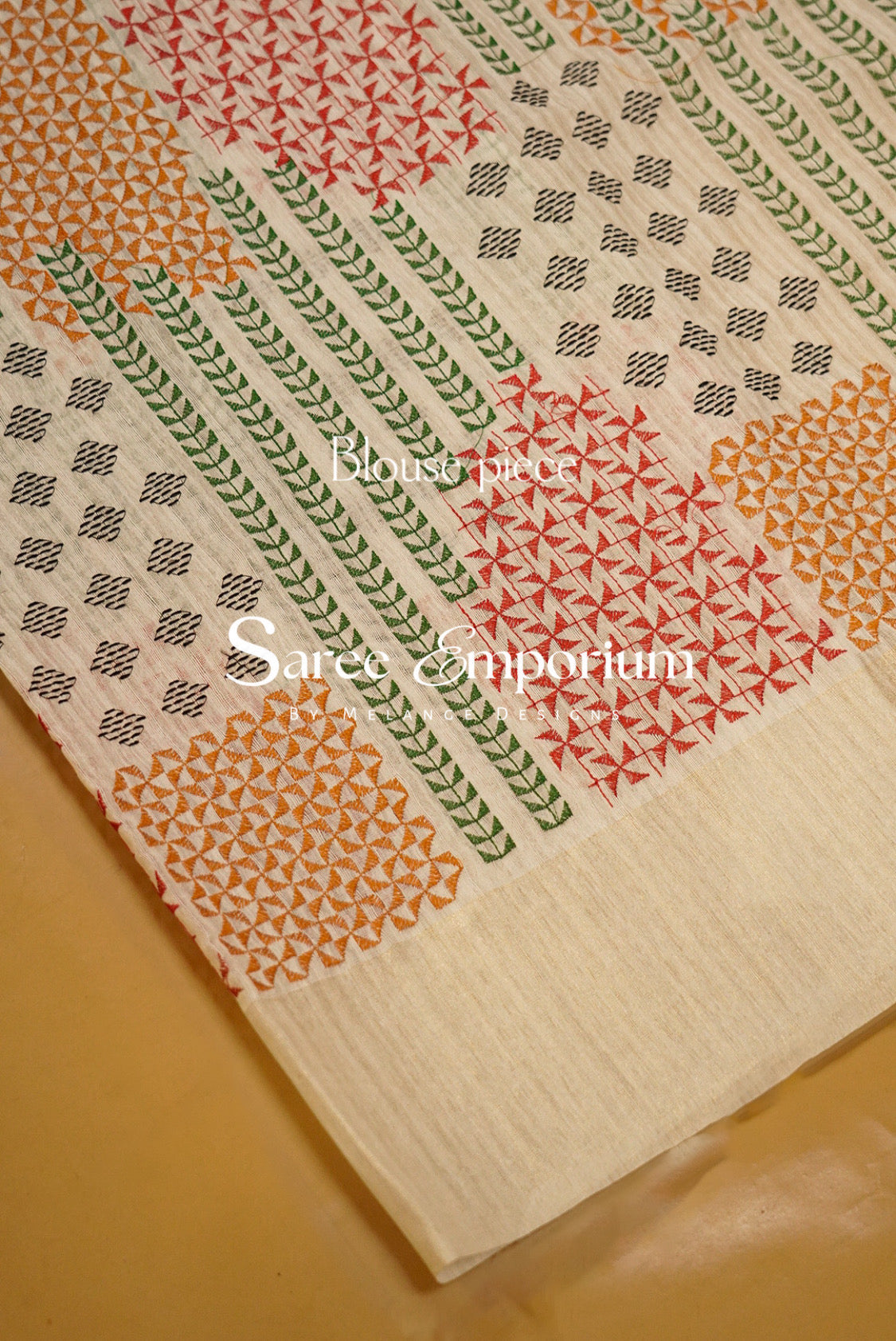 A close-up of a patterned textile laid on a warm beige surface. The fabric has a off white base with repeating geometric and floral motifs in orange, red, green, and black, plus a solid cream border.