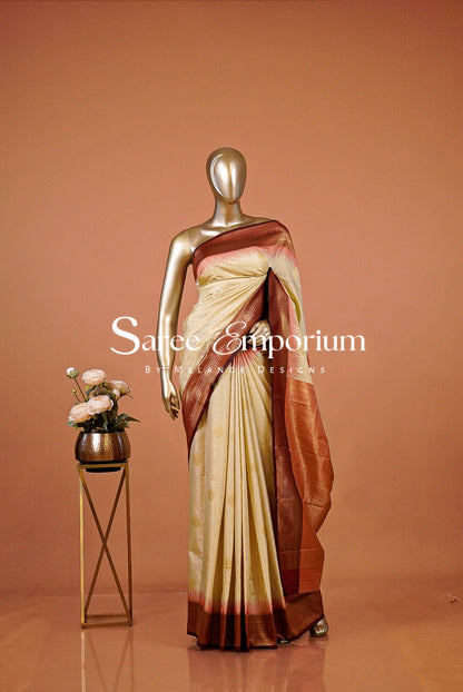 A graceful Semi Silk Saree in a soft ivory-cream tone, beautifully accented with a rich maroon and antique-gold zari border. Delicate golden woven motifs are scattered across the body, while subtle coral-red detailing along the border adds a refined traditional touch. The intricately woven pallu features elegant geometric and floral zari patterns for a luxurious finish.