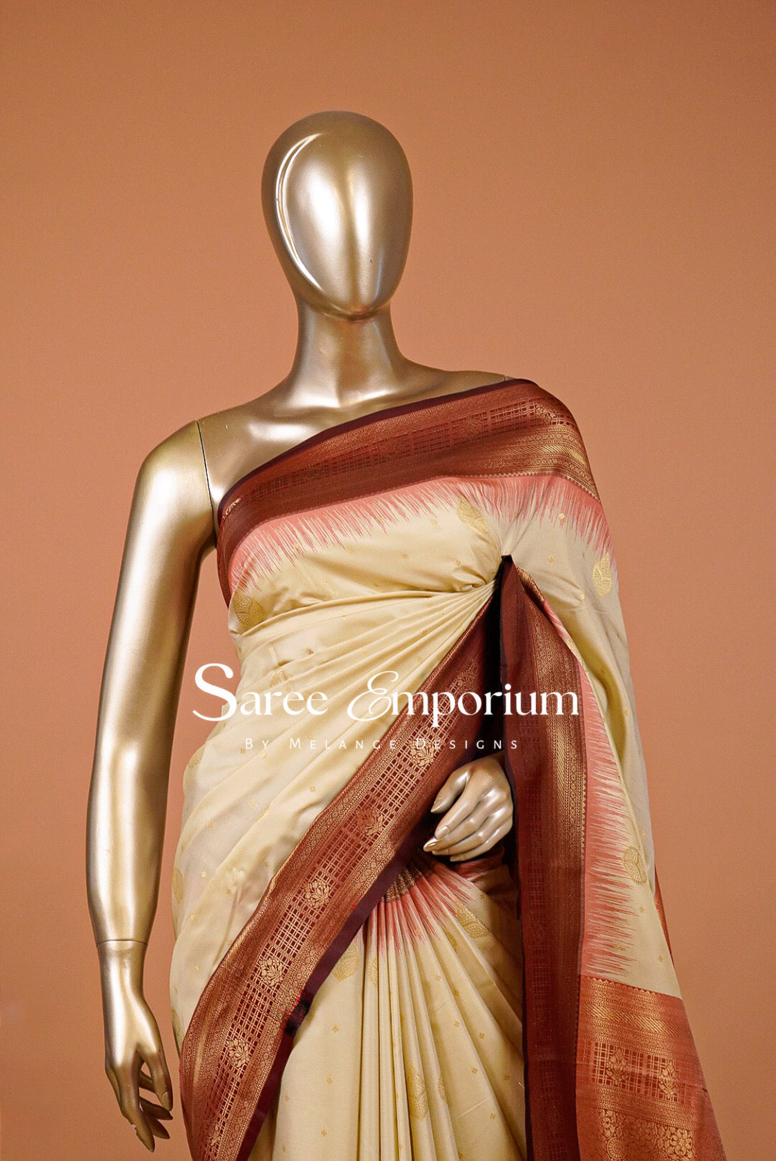 A graceful Semi Silk Saree in a soft ivory-cream tone, beautifully accented with a rich maroon and antique-gold zari border. Delicate golden woven motifs are scattered across the body, while subtle coral-red detailing along the border adds a refined traditional touch. The intricately woven pallu features elegant geometric and floral zari patterns for a luxurious finish.