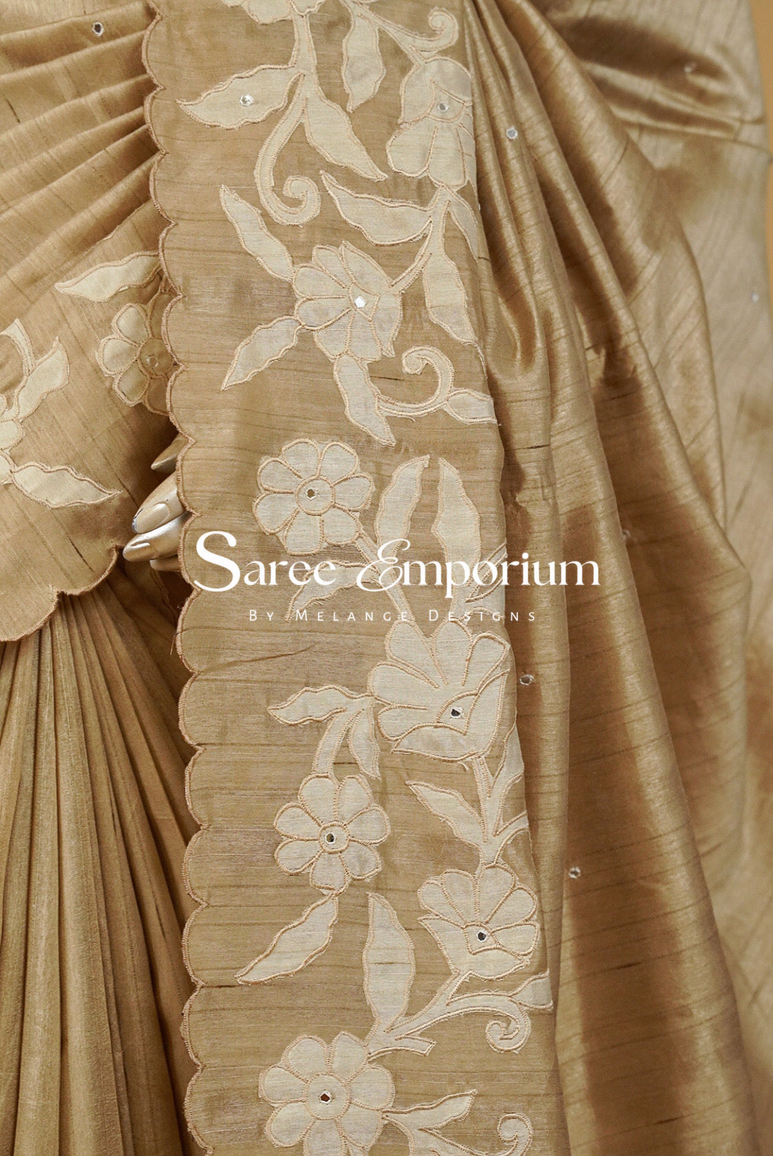 A close-up of a beige silk saree with a prominent border featuring cream floral embroidery and scalloped edges. The fabric has a subtle sheen.