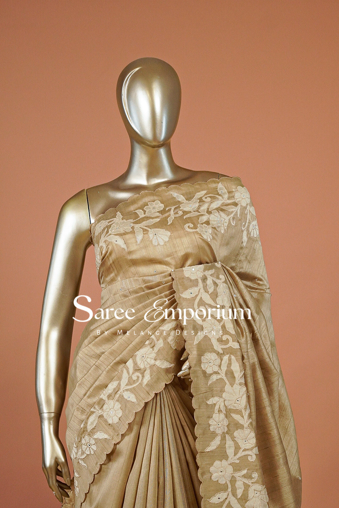 A metallic gold mannequin displays a champagne beige saree with white floral embroidery and scalloped borders.