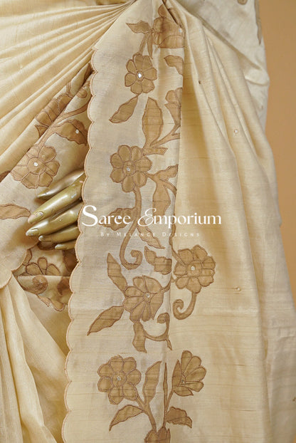 A close-up of a cream-colored silk saree with brown floral embroidery on the border, held by a hand with gold rings. 